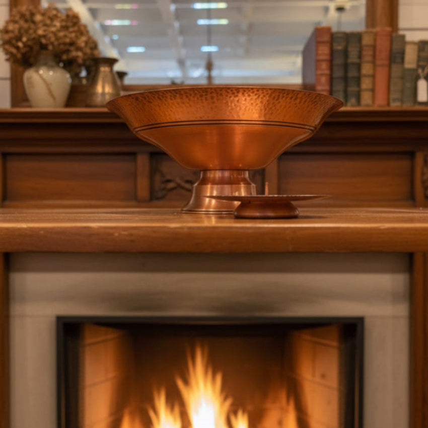 Side view of a copper pedestal bowl and matching pillar candleholder on a wooden mantle with a fire burning below