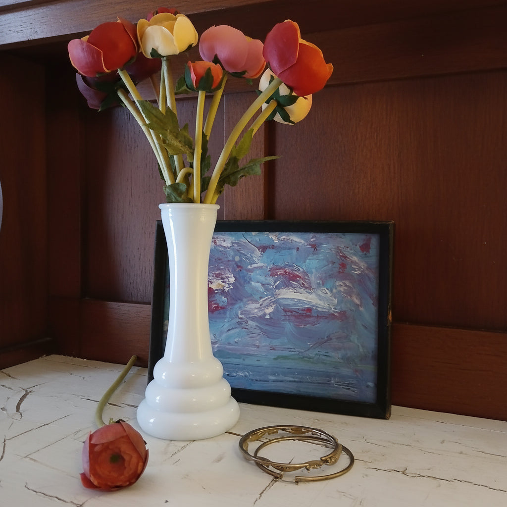 White glass vase with red and yellow flowers on a painted surface next to a framed painting.