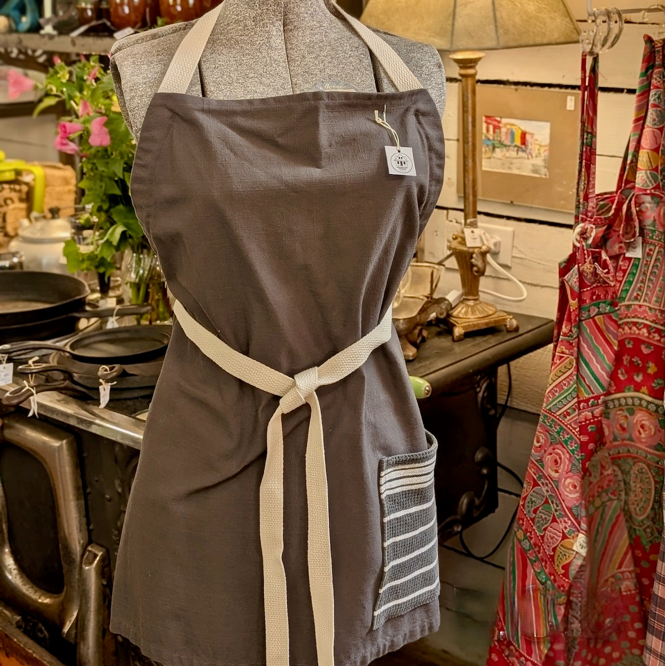 Grey linen apron with a white strap and grey and white pocket hanging on a mannequin in a kitchen setting.
