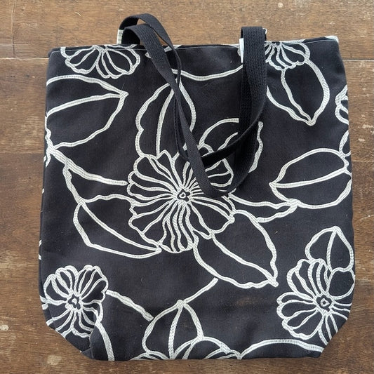 A black fabric tote bag with a vivid white geometric pattern shown on a wooden background