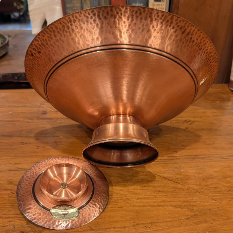A view of the bottom of a footed copper bowl and a matching candleholder on a wooden table with books in the background