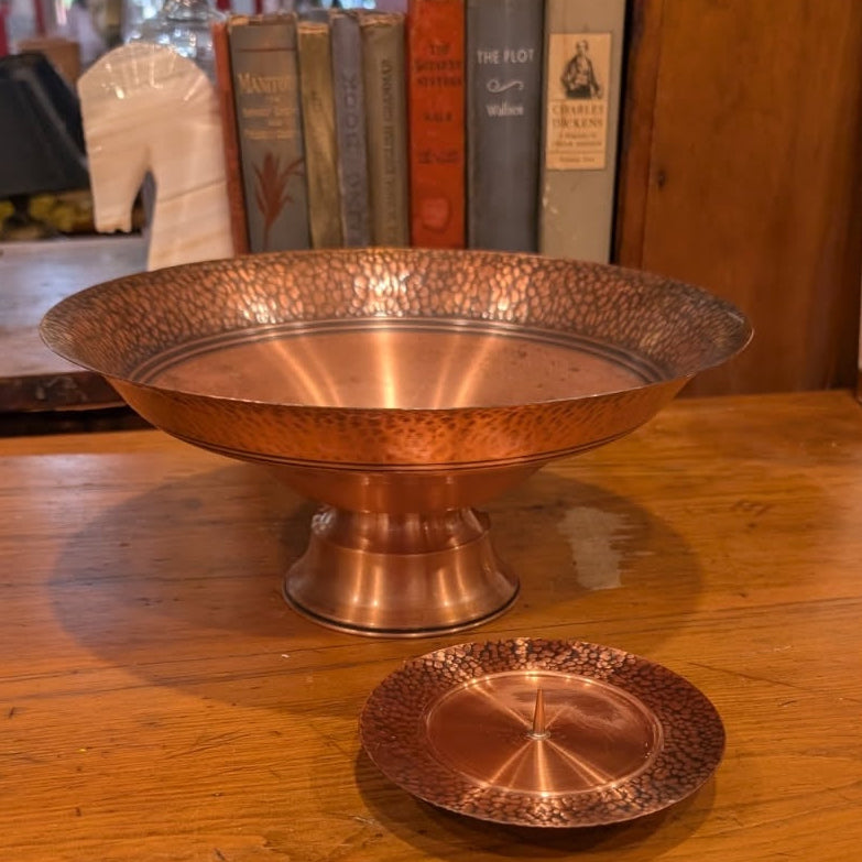 A copper pedestal bowl and matching copper pillar candleholder on a wooden surface with books in the background