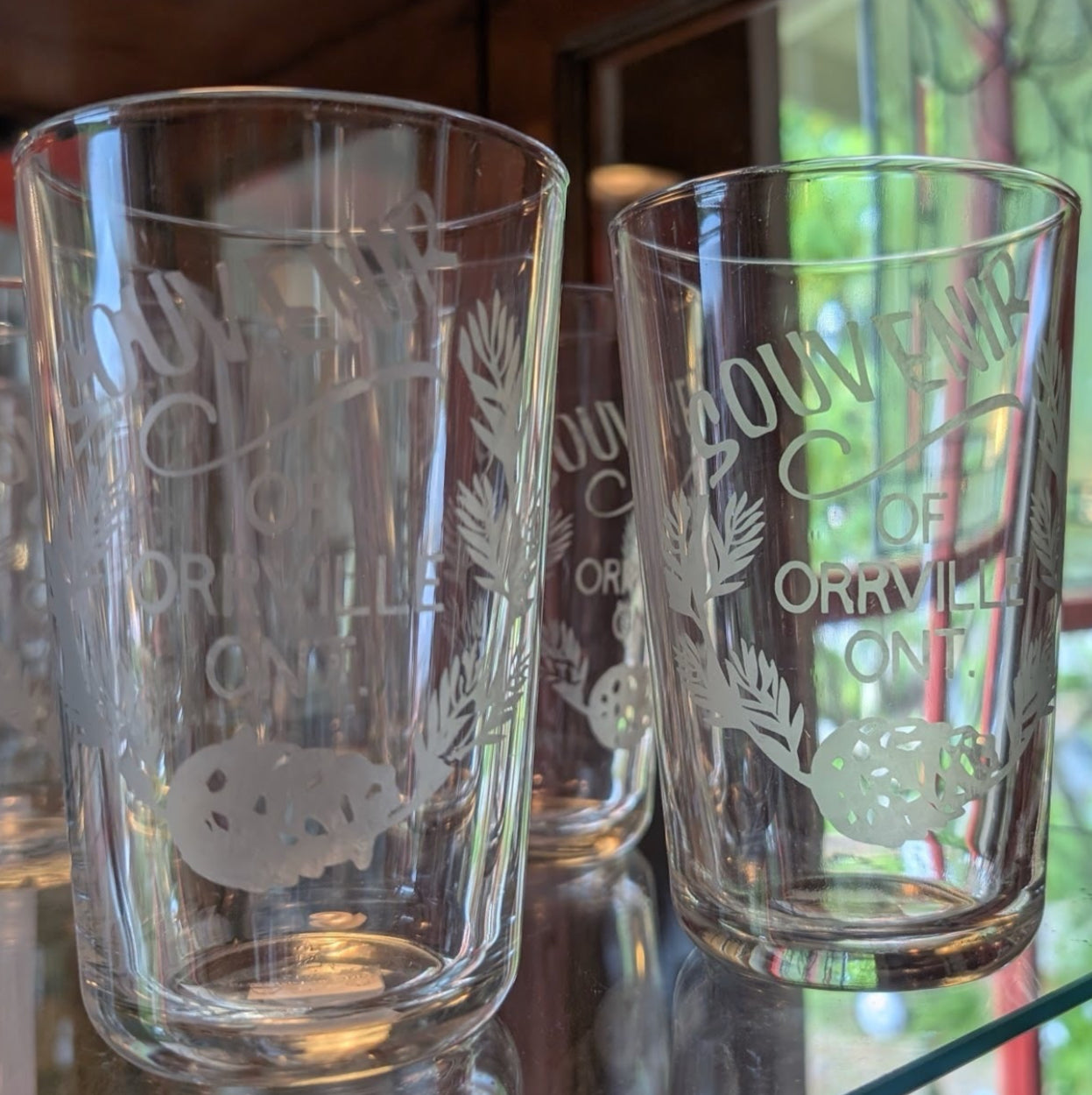 Three clear glasses with an edged design 'Souvenir of Orrville Ont.' on a shiny glass shelf