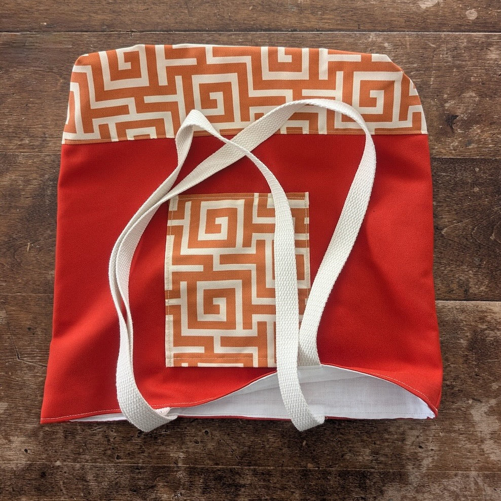 Hand-sewn orange tote bag with bronze geometric pattern on the pocket and the base showing a crisp white interior lining and white straps on a wooden surface