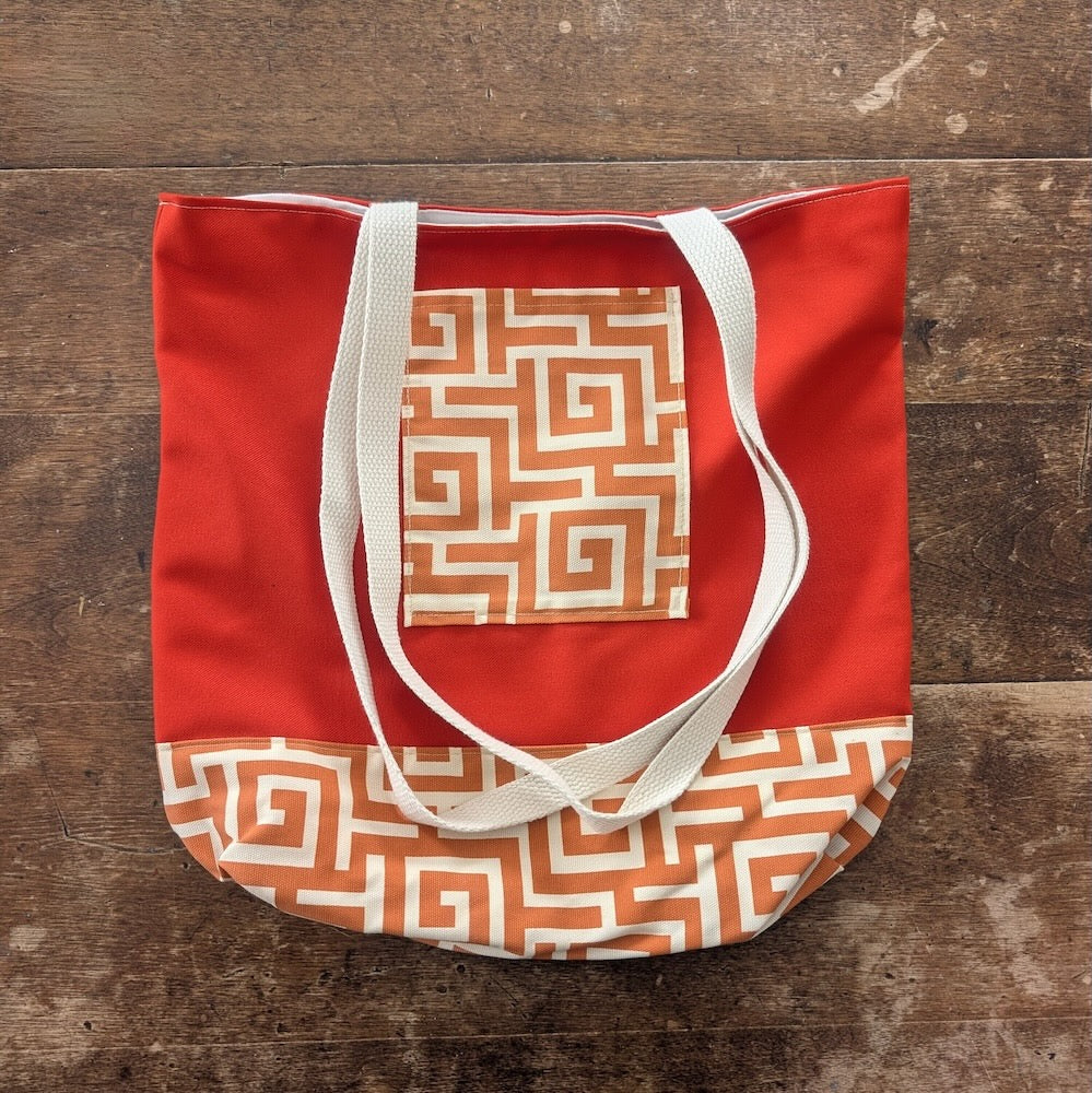 Hand-sewn orange tote bag with bronze-coloured geometric pattern on the pocket and the base with white straps on a wooden surface