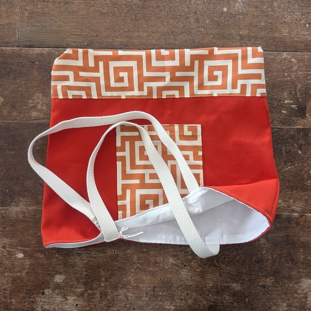 Hand-sewn orange tote bag with a bronze geometric pattern on the exterior pocket and the base, showing a  crisp white interior lining and white straps.