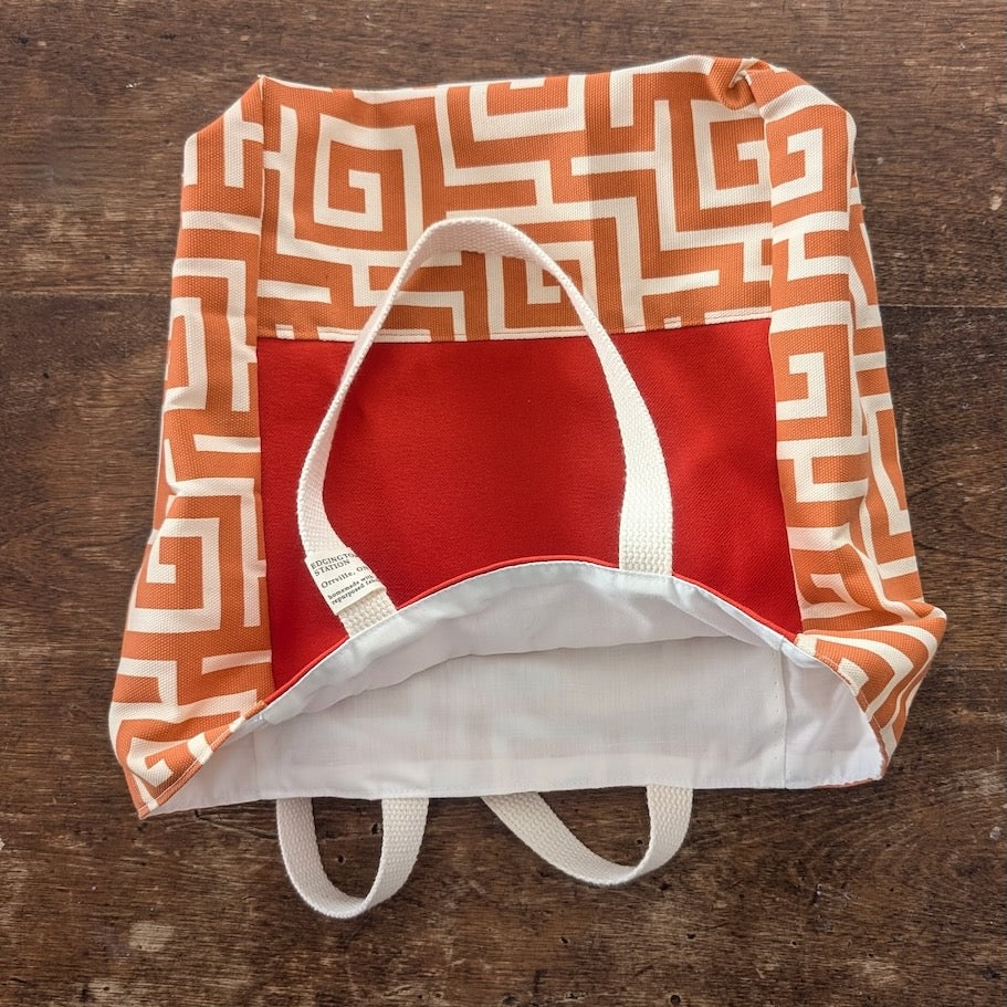 A small tote bag made with salvaged vintage fabric printed with an orange and white geometric pattern with a bright orange insert and white lining, shown on a wooden surface