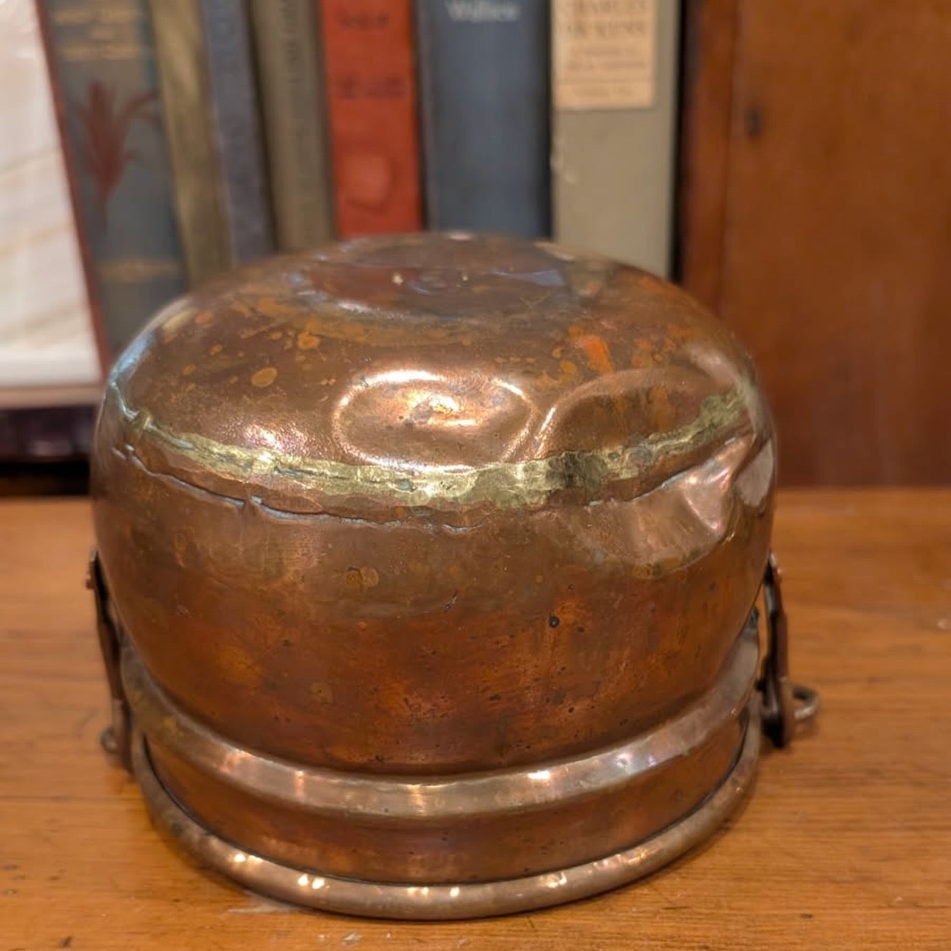 Vintage copper and brass container with a dented bottom on a wooden surface with books in the background