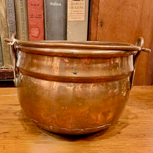 Battered copper and brass pot on a wooden surface with books in the background
