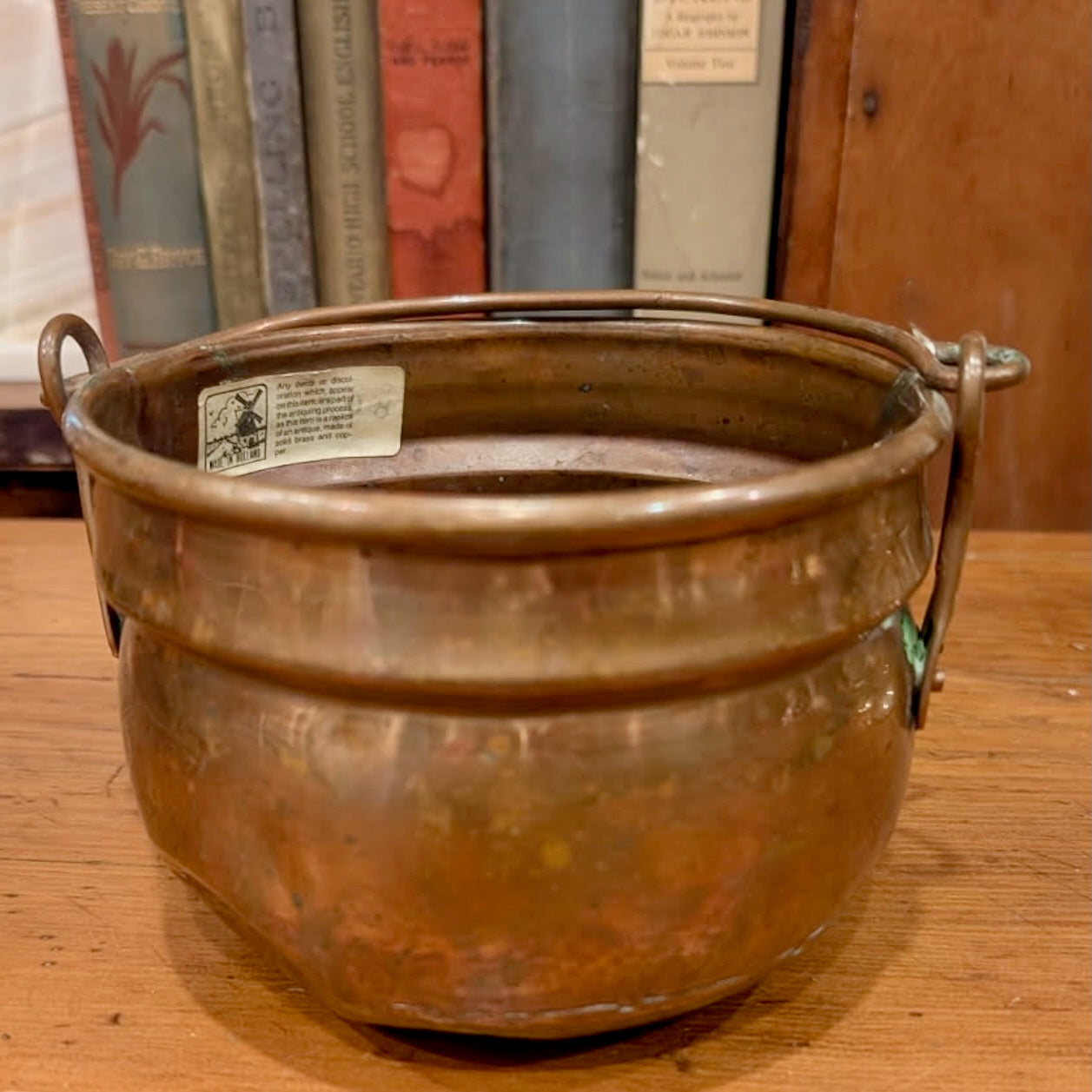 A vintage copper and brass pot with simple handles and a Holland sticker on a wooden surface, with books in the background