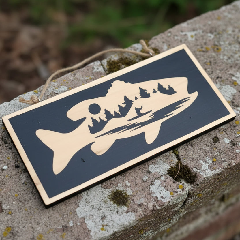 A closer view of a wooden wall plaque with painted image of a canoeist in the wilderness within the outline of a fish on a rustic stone sill.
