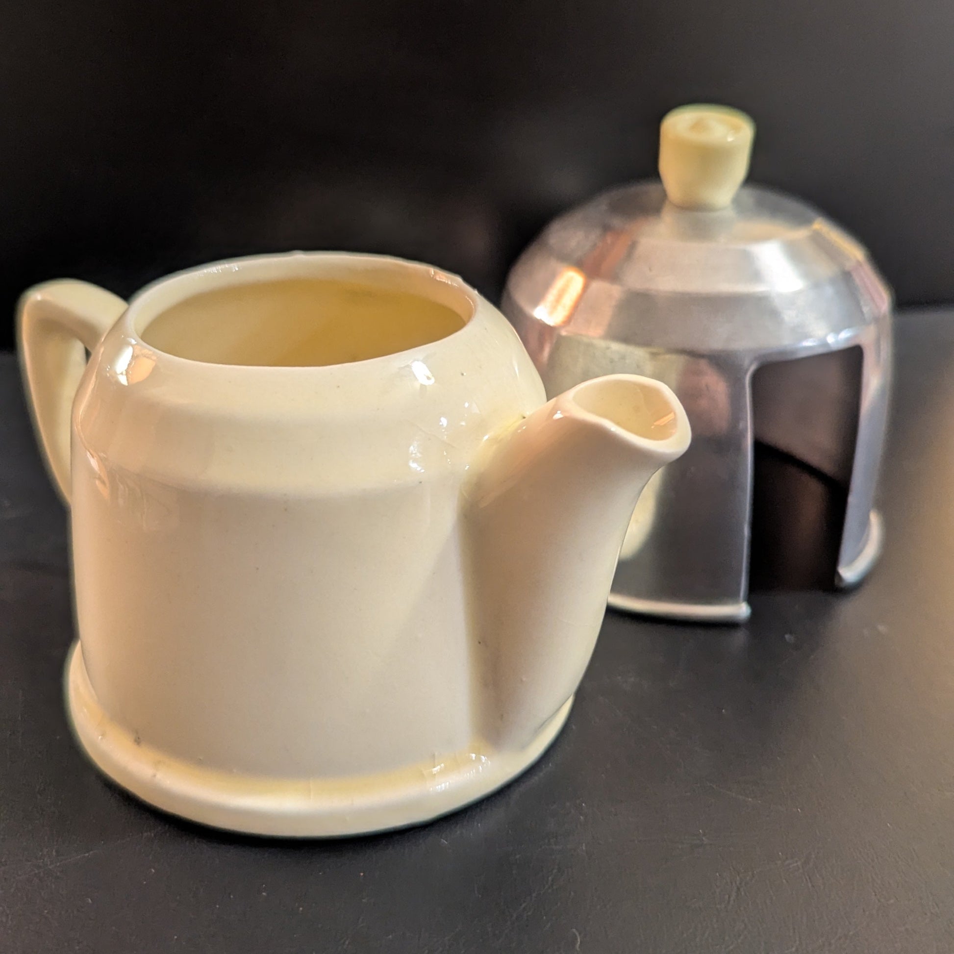 A small butter-yellow teapot with a shiny metal lid cozy against a dark background