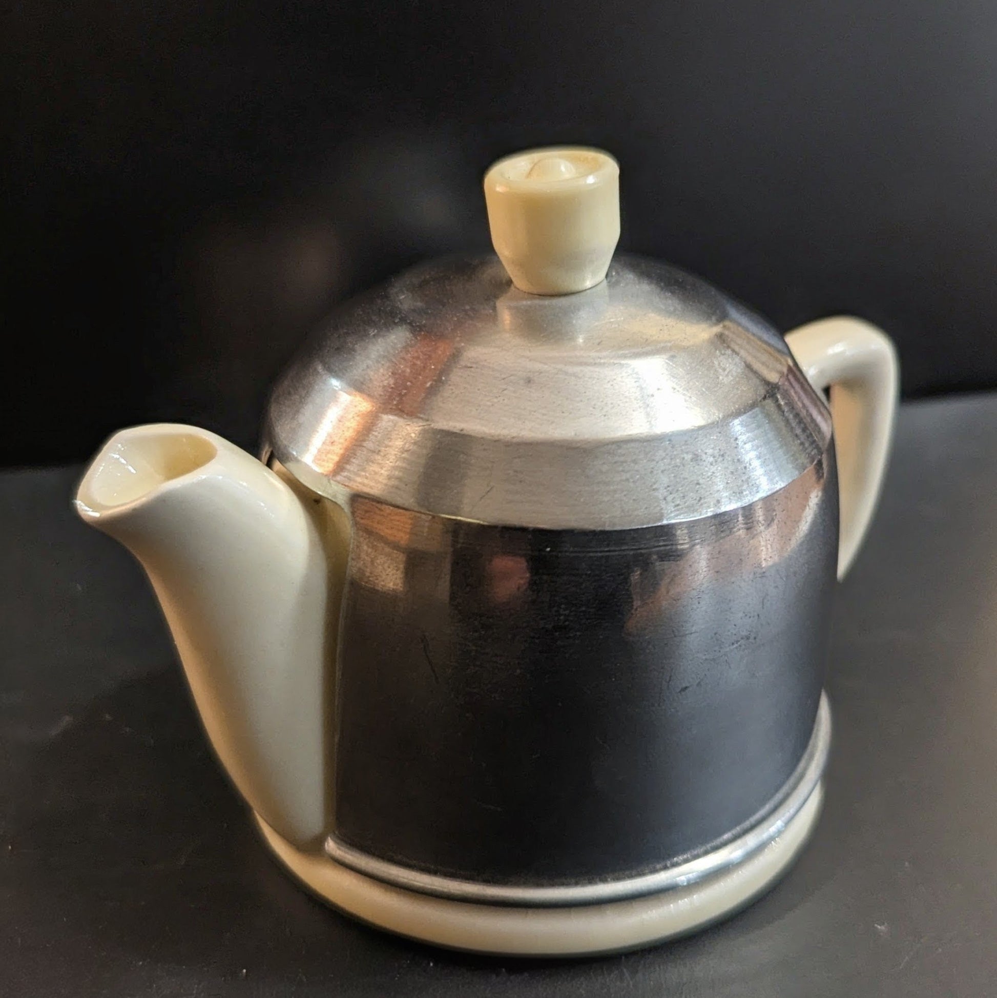 Small butter-yellow teapot with a metal lid that covers the entire pot except the spout and handle on a dark background