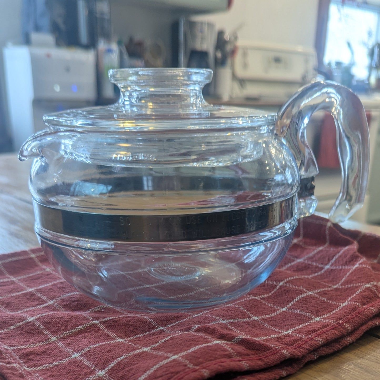 Clear glass teapot with glass handle on a red checkered towel on a wooden surface.