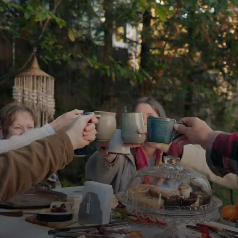 A small group toasts with mugs of warm drinks at a garden table set with snacks